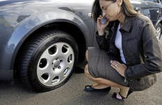Women on phone kneeling and looking at a flat tire on her car.
